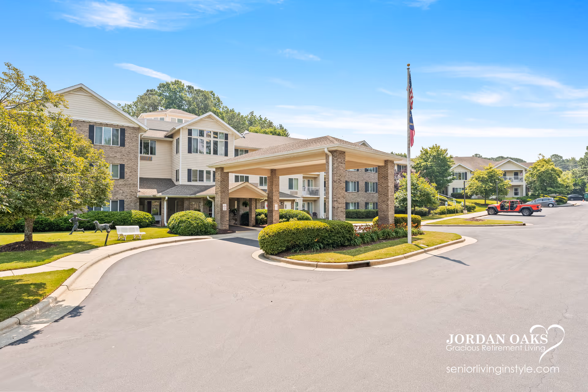 Exterior view of Jordan Oaks Gracious Retirement Living facility showing a large covered entrance with brick pillars, a flagpole with an American flag, well-maintained landscaping with bushes and trees, a curved driveway, and parked cars under a clear blue sky.