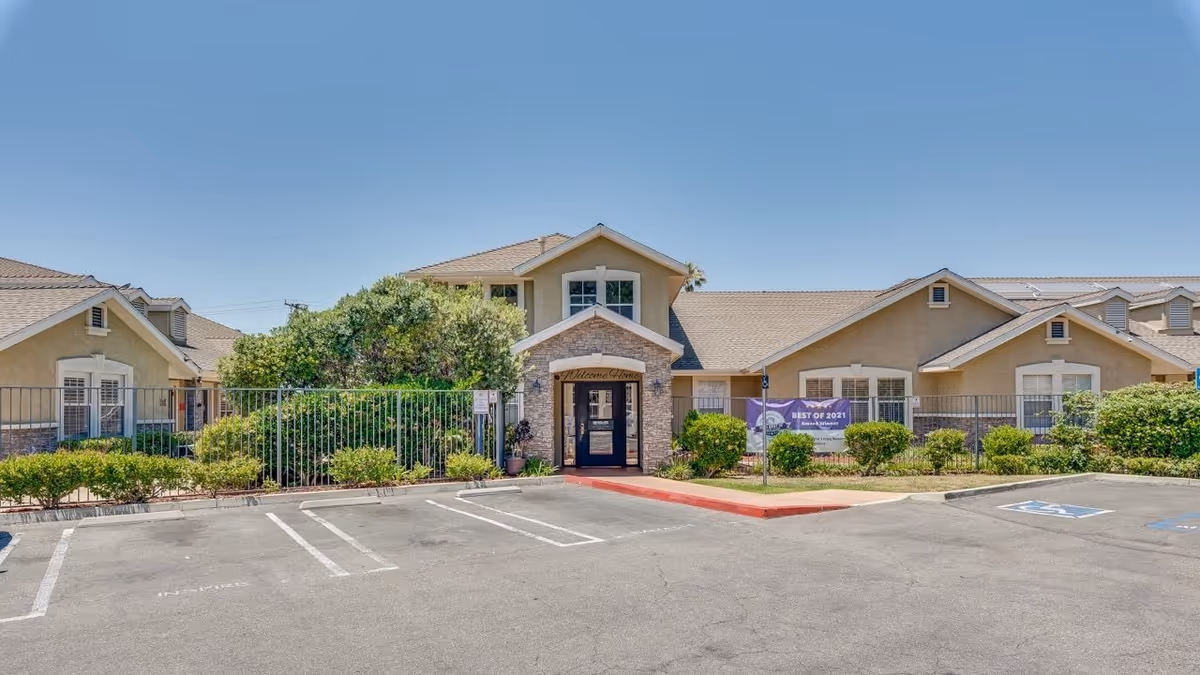 Exterior view of Newport Mesa Senior Living facility showing a single-story building with beige walls, a stone entrance, and a parking lot with marked spaces including handicapped spots. There are bushes and greenery around the building under a clear blue sky.