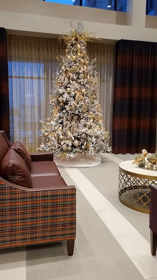 A decorated Christmas tree with gold and white ornaments stands in a well-lit room with large windows covered by curtains. In the foreground, there is a plaid-patterned sofa with brown cushions and a round coffee table with a decorative centerpiece.