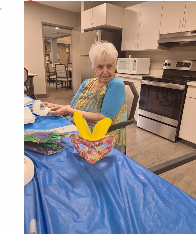 An elderly woman with white hair sits at a table covered with a blue plastic tablecloth in a kitchen area. She is holding a white paper mask and looking at the camera. On the table in front of her is a colorful mask decorated with yellow feathers. The kitchen has white cabinets, a stainless steel oven, microwave, and refrigerator. In the background, there is a dining area with chairs and tables.