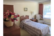 Single-occupancy bedroom with a twin bed and floral bedspread, dresser with a flower arrangement, nightstand, and an armchair by a window.