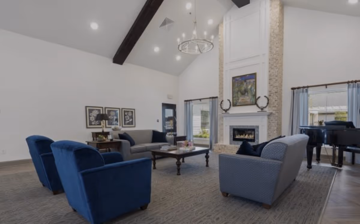 A spacious living room with high vaulted ceilings featuring a dark wooden beam and a modern chandelier. The room includes a fireplace with a stone surround and a painting above it, two blue armchairs, a gray sofa, a gray loveseat, a wooden coffee table, and a grand piano near a window with light blue curtains.