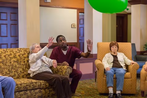 An elderly woman sitting on a floral-patterned couch reaching up towards a green balloon, assisted by a young man in a burgundy uniform. Another elderly woman sits nearby in a beige armchair, watching the activity. The setting appears to be a common area in a senior living facility with warm-colored walls and carpeted floor.