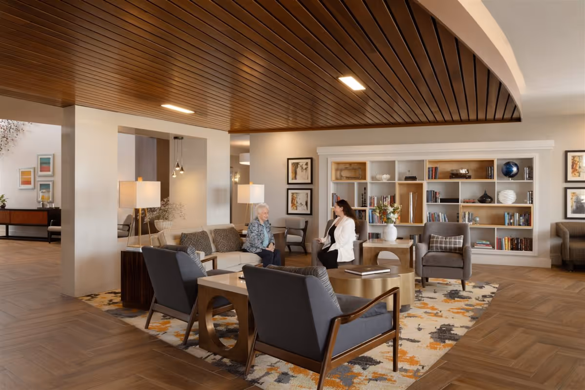 A cozy living room area in a senior living facility with two women sitting and conversing on a sofa and armchair. The room features a wooden ceiling, a patterned rug, modern furniture including armchairs and a coffee table, and a large built-in bookshelf filled with books and decorative items.