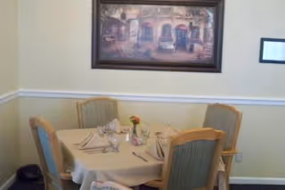 A small dining area with a round table covered with a beige tablecloth, set with folded napkins, glasses, and silverware. Four upholstered chairs with wooden frames surround the table. A framed painting depicting an outdoor scene hangs on the light-colored wall above the table.