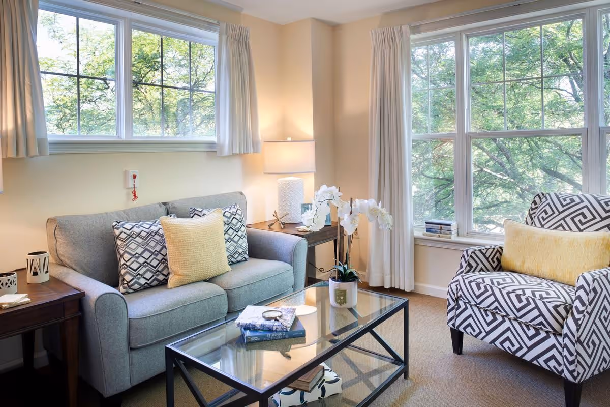 Bright living room with a sofa, patterned armchair, glass coffee table, and large windows showing trees outside.