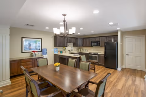 A well-lit kitchen and dining area featuring a wooden dining table with six chairs, a modern kitchen with dark wood cabinets, black refrigerator, stove, microwave, and a countertop with a sink. The room has wood flooring, a chandelier above the dining table, and a framed picture on the wall above a sideboard with a water dispenser and decorative flowers.