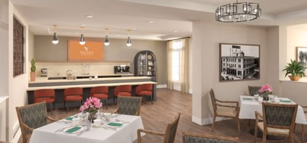 A bright and spacious dining area in a senior living facility featuring tables set with white tablecloths, green napkins, and pink floral centerpieces. The room has wooden flooring, several chairs around the tables, and a counter with red bar stools. Pendant lights hang above the counter, and there is a framed black and white photograph on the wall along with a potted plant near a window with sheer curtains.