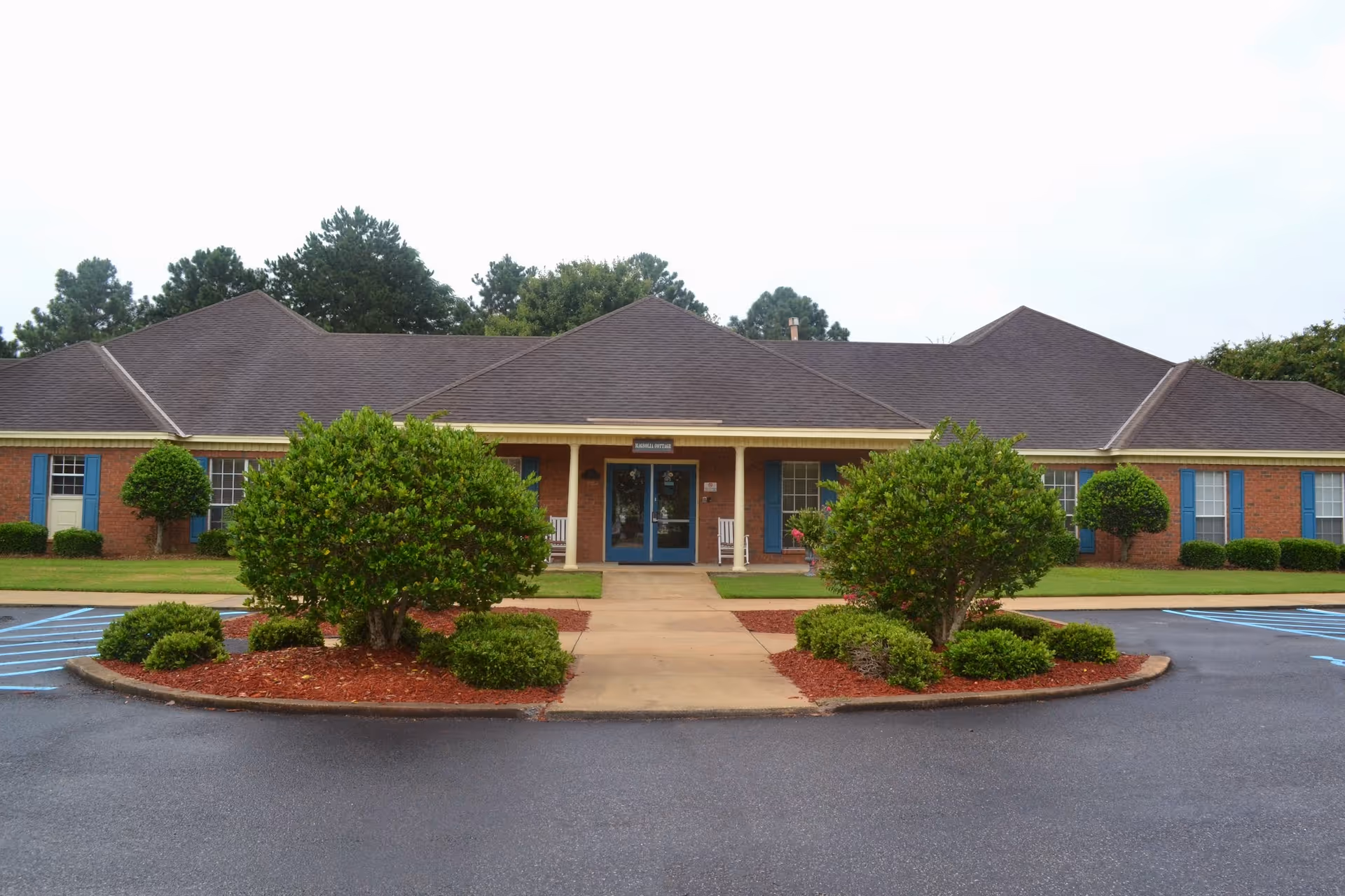Front exterior view of a single-story brick building with a dark shingled roof, blue window shutters, and a central entrance with double doors. The entrance is flanked by two white rocking chairs and surrounded by neatly trimmed bushes and landscaped areas with red mulch. The parking lot in front has marked spaces, including accessible parking spots.