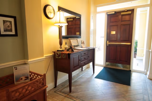 Entrance area of a facility with a wooden door, a dark wooden console table with a lamp, framed pictures, and a wall clock. There is a green floor mat in front of the door and a small wooden bench with a framed photo on it to the left.