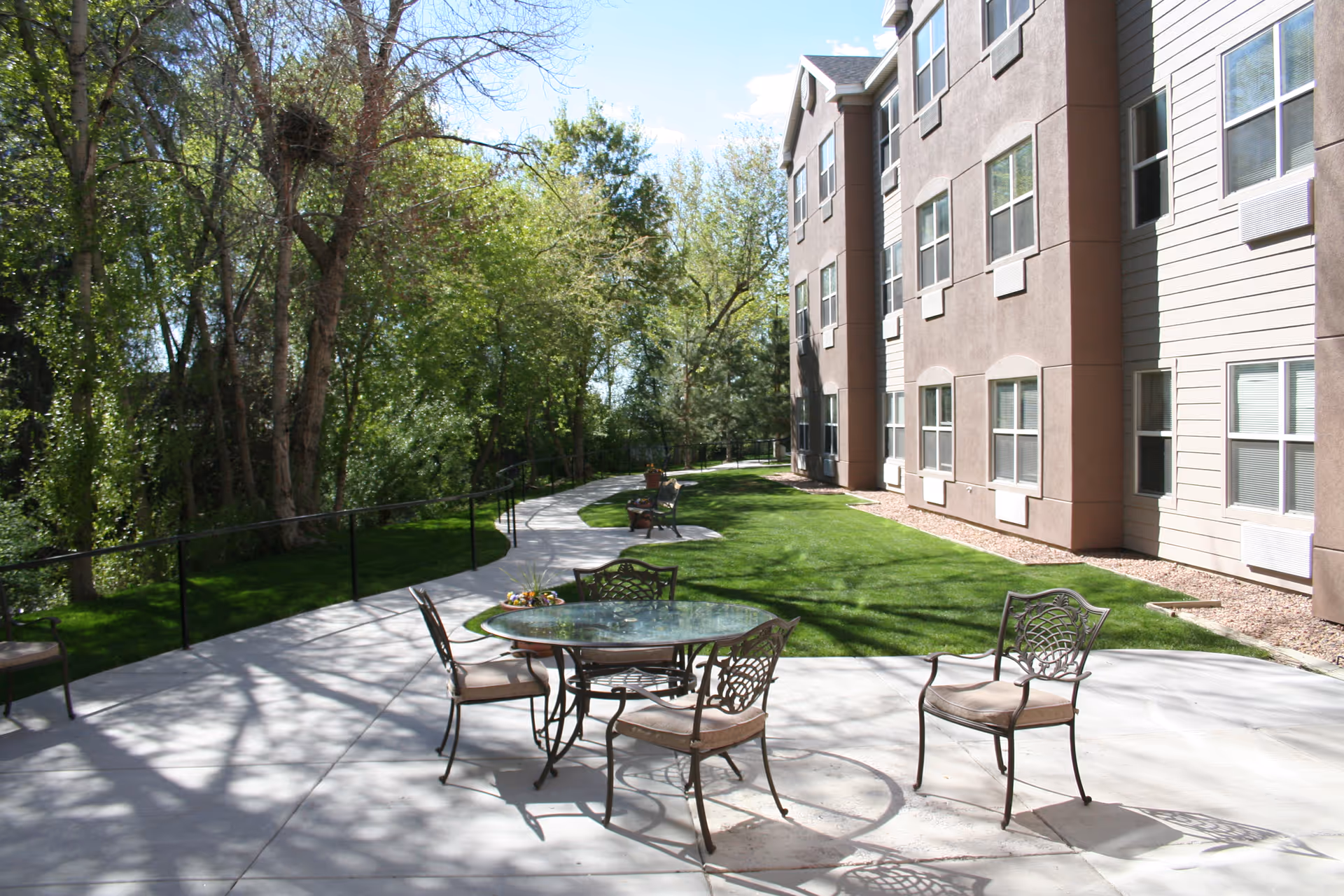 Outdoor patio courtyard with metal table and chairs, a winding walkway, lawn and trees beside a multi-story residential building.