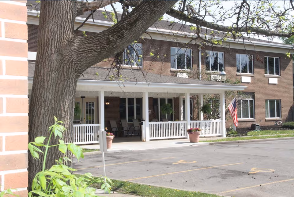 Exterior view of Orchard Brooke Living Center showing a two-story brick building with multiple windows, a covered porch with white railings, several chairs, potted plants, and an American flag near the entrance. A large tree is in the foreground and there is a parking area with handicap parking spaces.