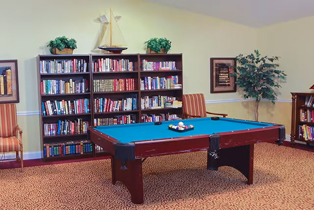 A cozy room with a pool table in the center, surrounded by bookshelves filled with books. There are two striped armchairs on either side of the bookshelves, framed artwork on the walls, and a decorative sailboat on top of the bookshelf. A potted plant is placed in the corner, and the room has patterned carpet flooring.