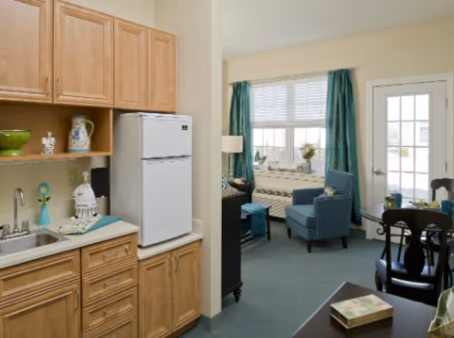 Small apartment-style living area showing a kitchenette with wood cabinets and a refrigerator opening into a seating area with teal curtains, armchairs, and a dining table by a glass door.