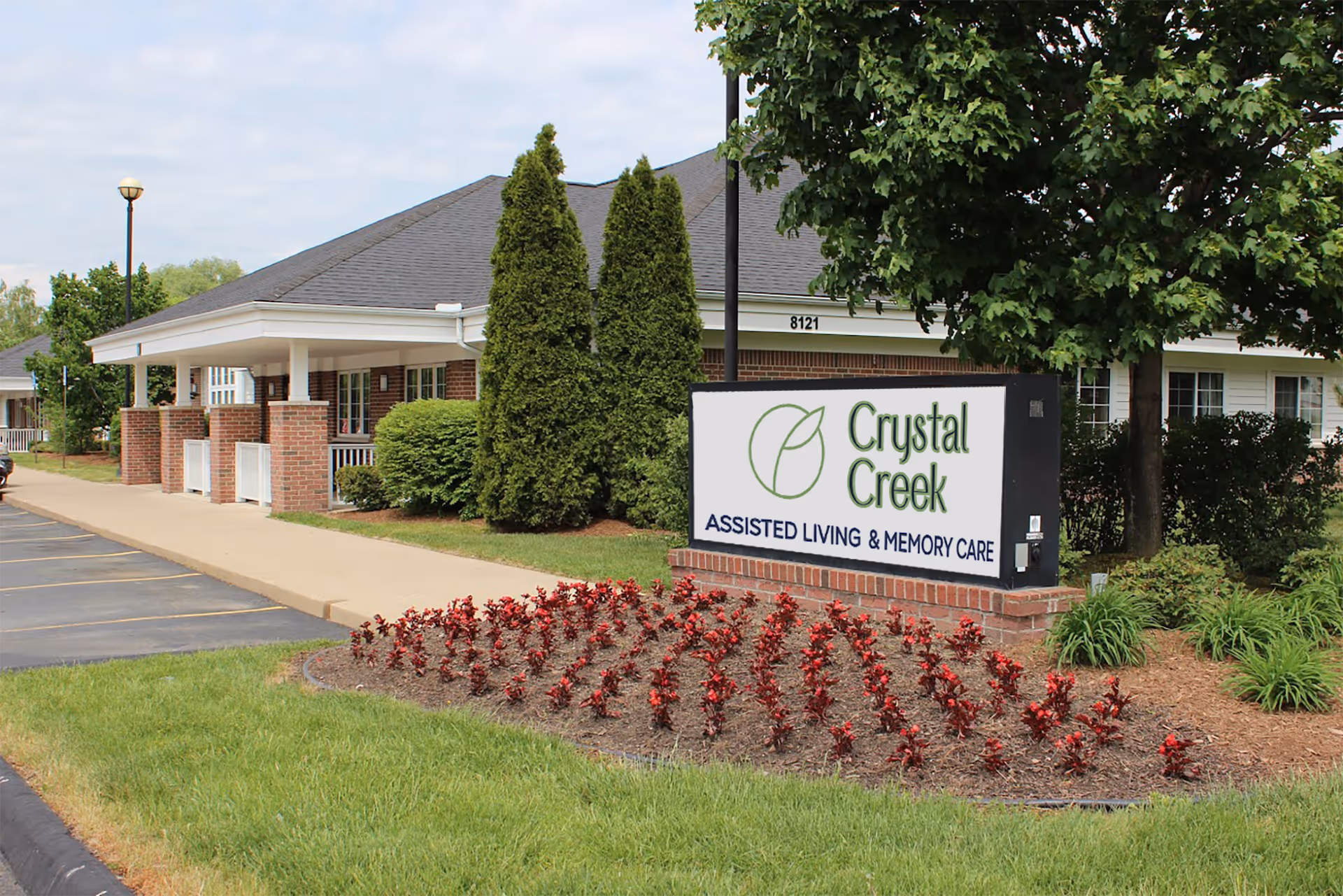Exterior view of Crystal Creek Assisted Living and Memory Care facility showing a brick building with a covered porch, landscaped garden with red flowers, green bushes, and trees, and a sign with the facility's name in front.