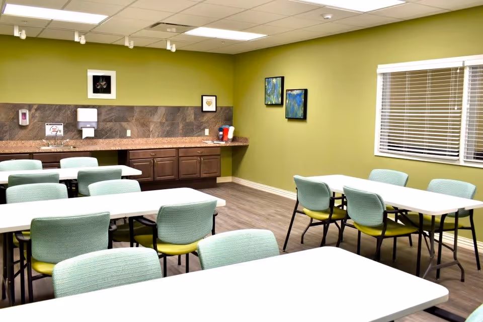 A brightly lit room with green walls and several white tables surrounded by light green chairs. The room has a countertop with cabinets, a sink, a soap dispenser, and framed artwork on the walls. There is a window with blinds on the right side.