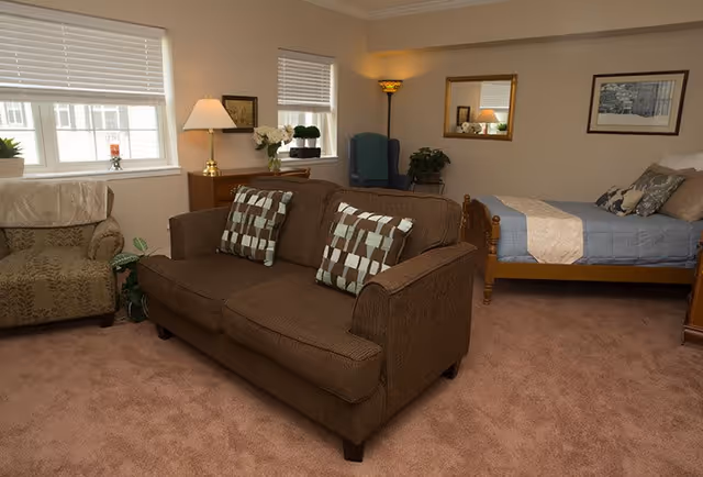 A cozy senior living room and bedroom area with a brown couch featuring two patterned pillows, an armchair, a wooden bed with blue bedding, a wooden dresser with a lamp and flowers, a tall floor lamp, and framed pictures on beige walls.