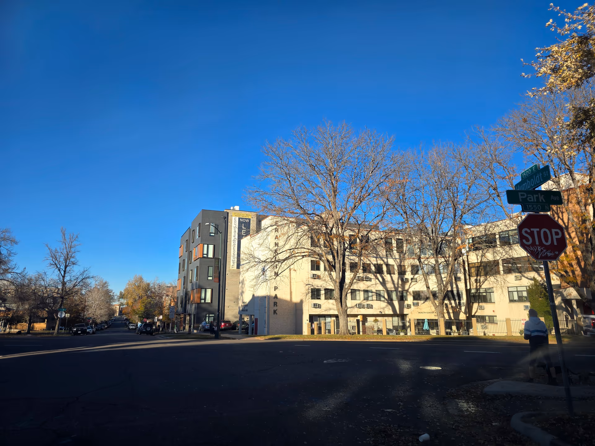 Exterior view of the Health Center at Franklin Park building on a clear day with a bright blue sky. The building is a multi-story structure with several windows and air conditioning units. Leafless trees are in front of the building, and a street intersection with stop signs and street name signs for Humboldt St and Park Ave is visible. A person is walking on the sidewalk near the stop sign.