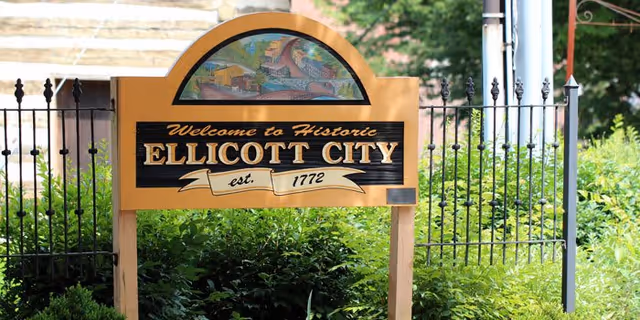 A wooden sign with a decorative top panel displaying a colorful illustration, reading 'Welcome to Historic Ellicott City est. 1772', surrounded by green bushes and black iron fencing in the background.