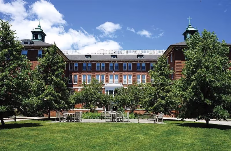 A large brick building with multiple windows and two cupolas on the roof, surrounded by green trees and a well-maintained lawn with outdoor benches and tables under a partly cloudy blue sky.