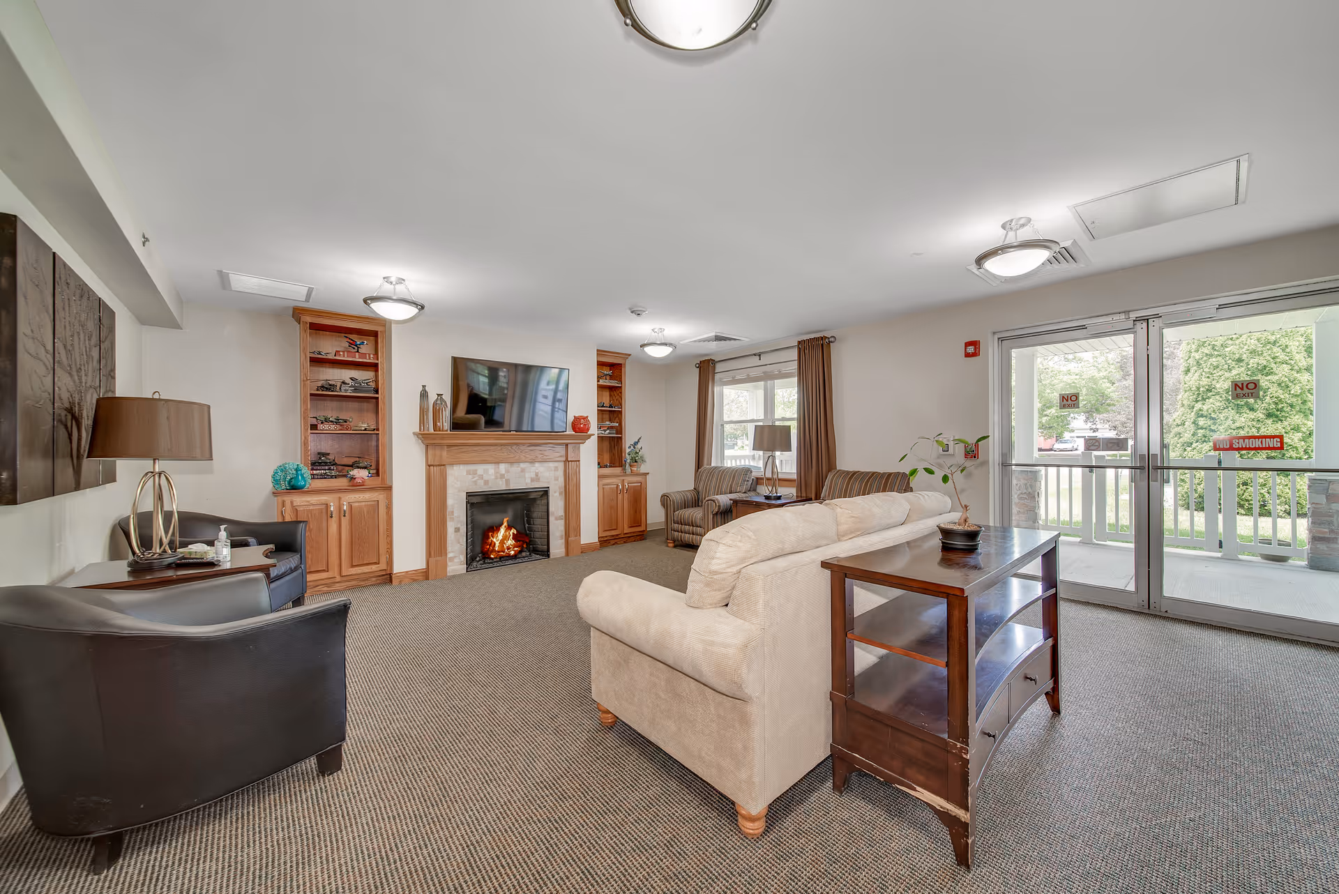 A cozy living room area in a senior living facility with a beige sofa, two striped armchairs, and two black armchairs arranged around a fireplace with a mounted TV above it. Wooden built-in shelves flank the fireplace, and large windows with brown curtains let in natural light. A wooden side table with a plant is placed behind the sofa, and glass doors lead to an outdoor area with greenery visible outside.