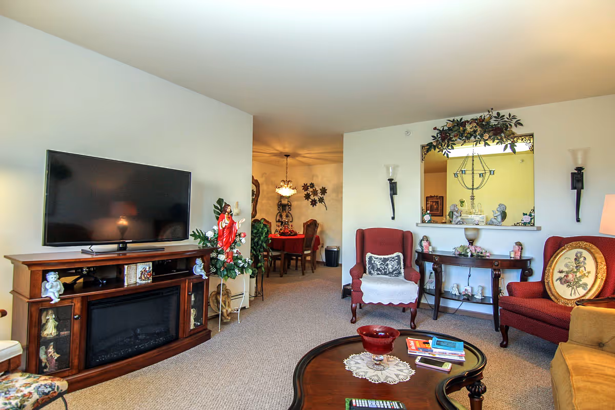 A cozy living room in Villa Ciera Senior Apartments featuring a flat-screen TV on a wooden stand with decorative figurines, two red upholstered armchairs, a wooden coffee table with books and a remote control, and a console table with decorative items. The room opens into a dining area with a chandelier and a table covered with a red tablecloth.