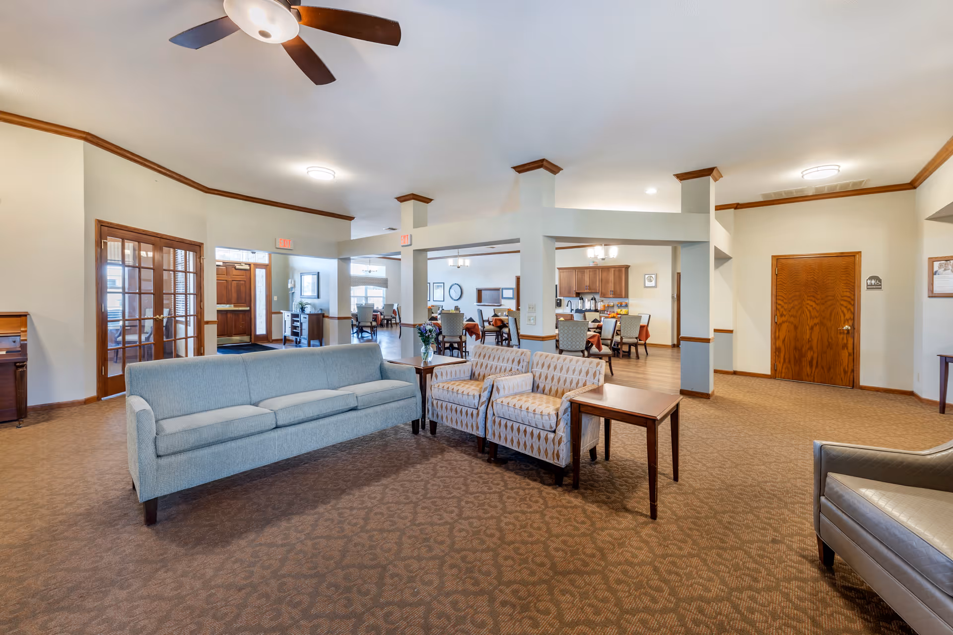 A spacious senior living facility common area with a light blue sofa, two patterned armchairs, and a small wooden table. The room features beige walls with wooden trim, a ceiling fan, and carpeted floor. In the background, there is a dining area with tables and chairs, a kitchenette with cabinets, and multiple exit signs above doors.