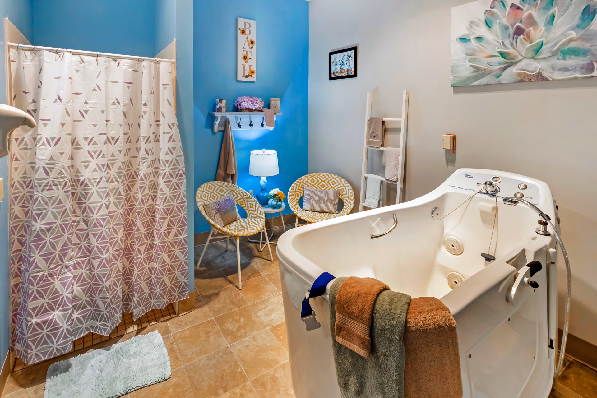 Bright bathroom with a walk-in bathtub, a shower with a patterned curtain, and two chairs with a small table against a blue accent wall.