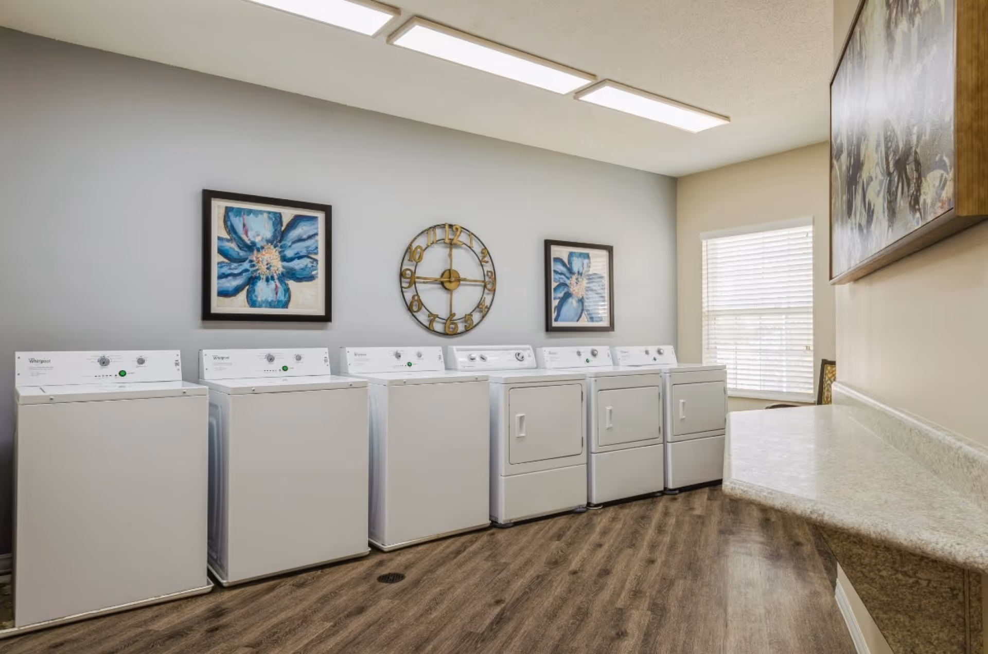 Laundry room with three washing machines and three dryers lined up against a light gray wall. Above the machines are two framed blue flower paintings and a decorative wall clock. The room has wood-style flooring, a window with blinds, and a countertop on the right side.