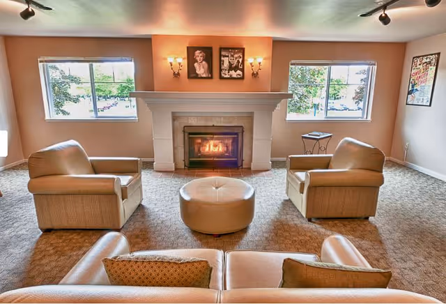 A cozy living room with a lit fireplace centered between two windows. The room features two beige armchairs facing the fireplace, a round beige ottoman in the middle, and a beige sofa with two cushions in the foreground. There are framed pictures above the fireplace and on the walls, and the room is carpeted.