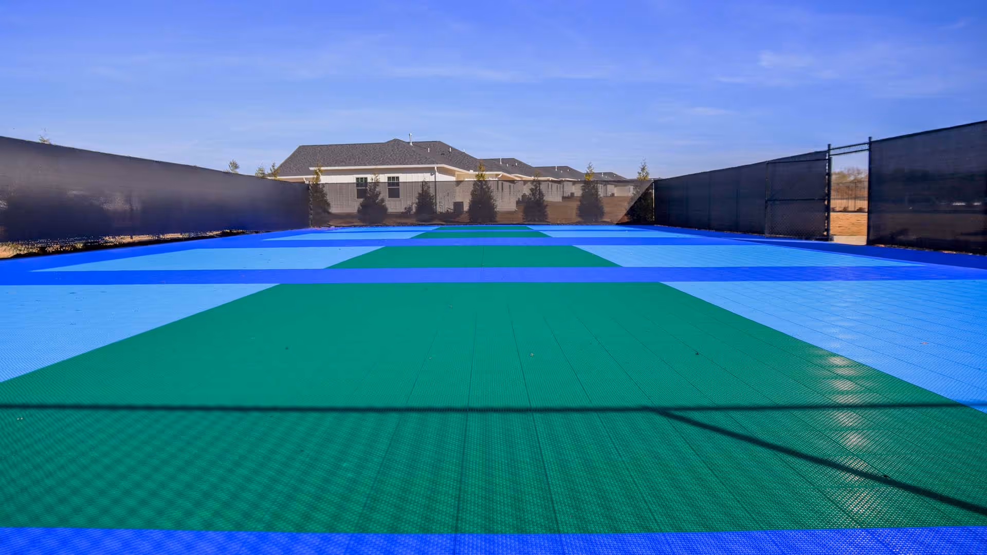 Outdoor fenced court with a blue and green checkered surface under a clear blue sky, with houses and trees visible in the background.
