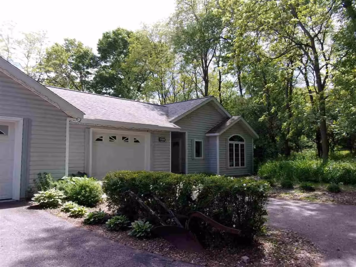 Single-story light-gray building with an attached garage, front shrubs and driveway, set among tall trees.