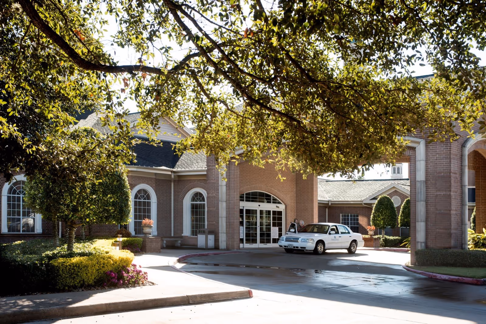 Exterior view of Buckner Parkway Place showing the entrance with a covered driveway. A white car is parked near the entrance with two people standing beside it. The building features brick walls and arched windows, surrounded by neatly trimmed bushes and trees with sunlight filtering through the leaves.