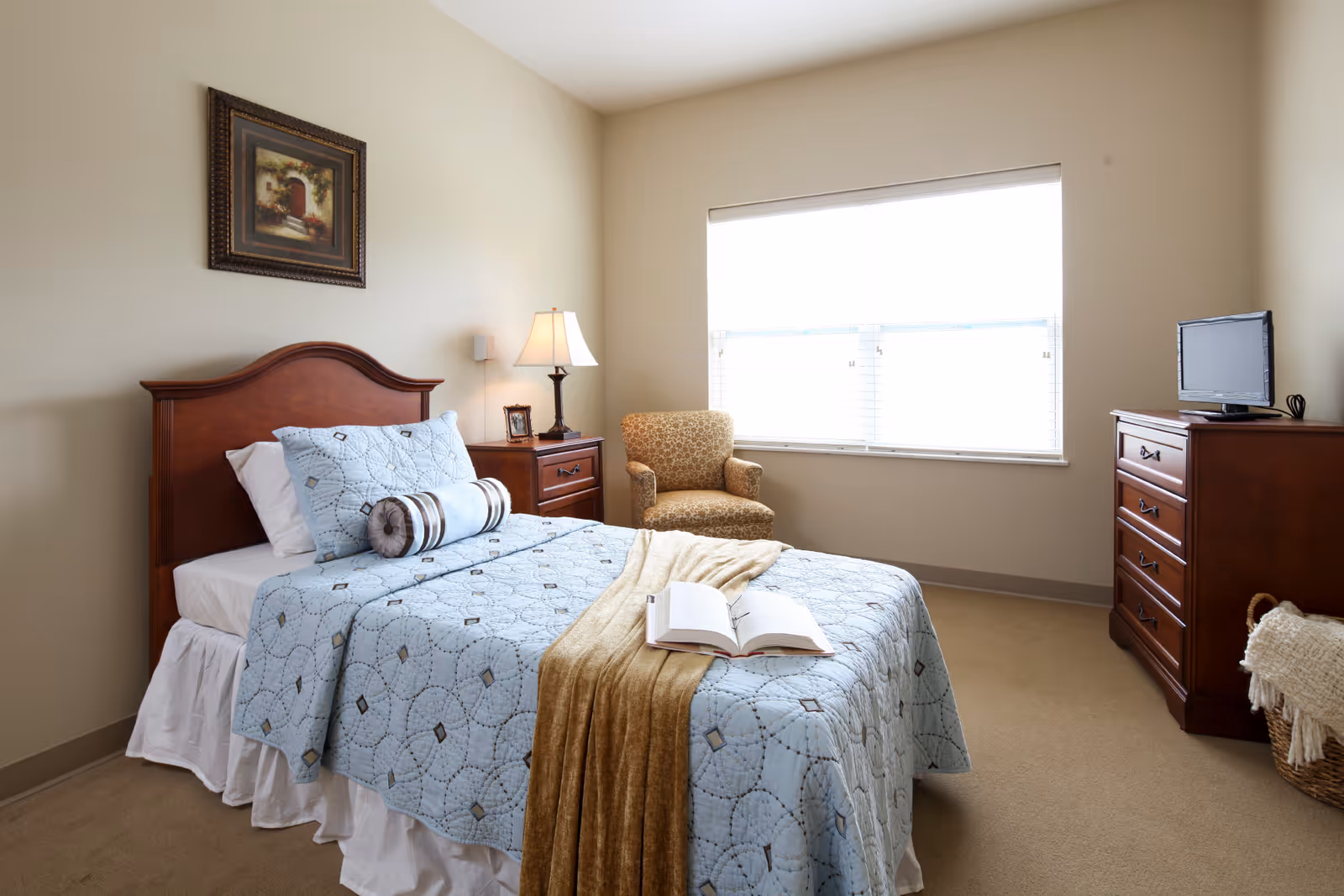 A neatly made single bed with a light blue patterned quilt and decorative pillows in a bedroom. A beige throw blanket and an open book are placed on the bed. Next to the bed is a wooden nightstand with a lamp and a small framed photo. A patterned armchair is positioned near a large window with white blinds. Across from the bed is a wooden dresser with a small flat-screen TV on top. A woven basket with a blanket is on the floor beside the dresser. The walls are painted a light beige color.
