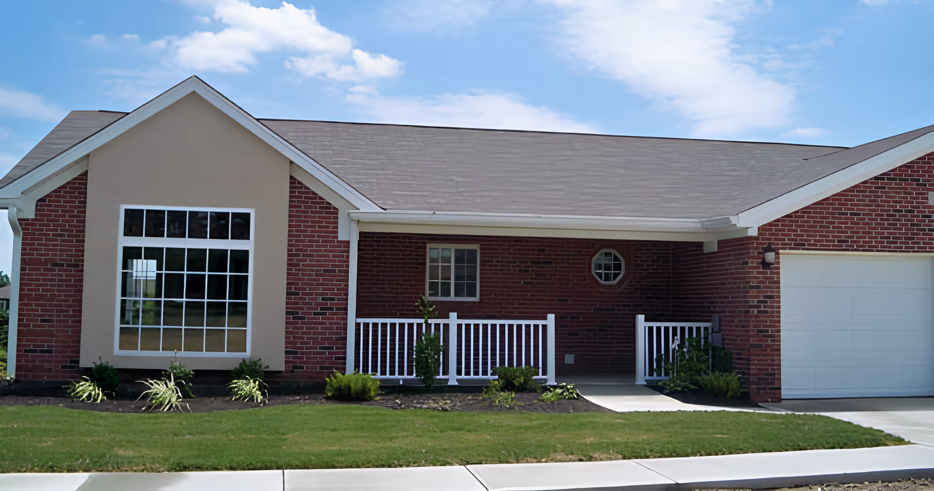 Front exterior of a single-story brick building with a porch, large window, attached garage, and lawn.