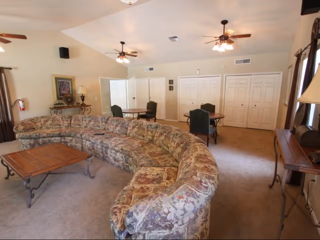 A spacious living room with a large curved floral-patterned sofa surrounding a wooden coffee table. The room has beige walls, carpeted floor, ceiling fans with lights, and several tables with chairs near white double-door closets. There are windows with curtains and decorative lamps on side tables.