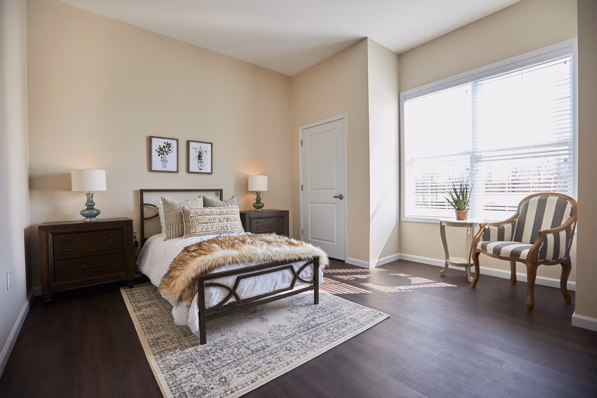 A bright bedroom with a single bed featuring white bedding and a fur throw. The bed is flanked by two dark wooden nightstands with matching lamps. Above the bed are two framed botanical prints. A patterned rug lies under the bed on dark wood flooring. To the right, a large window with white blinds lets in natural light, next to a small round table with a potted plant and a striped upholstered armchair.