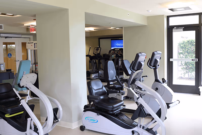 A bright interior fitness room with several recumbent exercise bikes, cardio machines, mirrors, and a glass exit door.