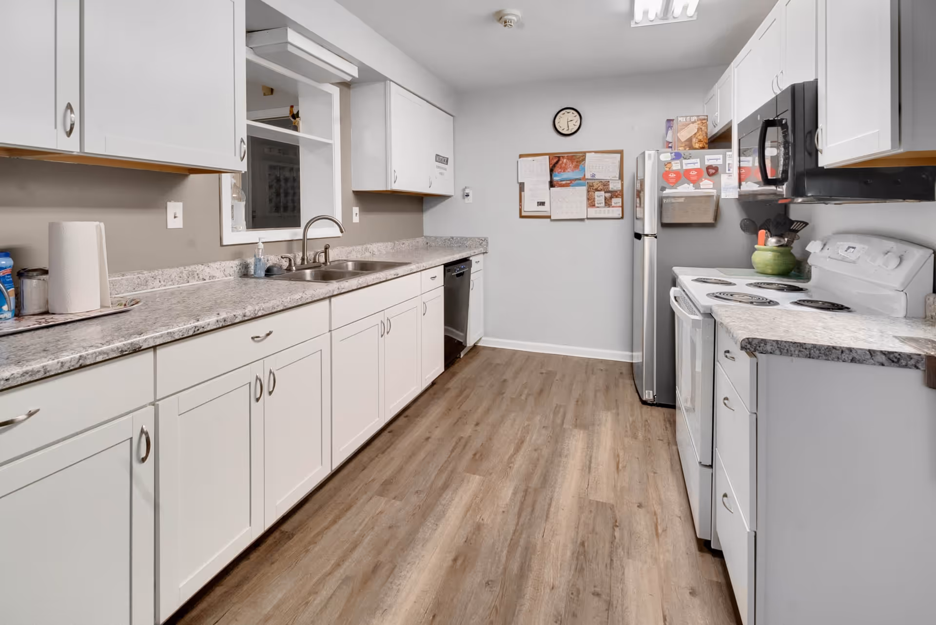 A clean, modern kitchen with white cabinets, a marble-patterned countertop, a double sink, a dishwasher, a white electric stove, a microwave, and a stainless steel refrigerator. The floor is wood-style laminate, and a bulletin board with papers and a clock is mounted on the far wall.