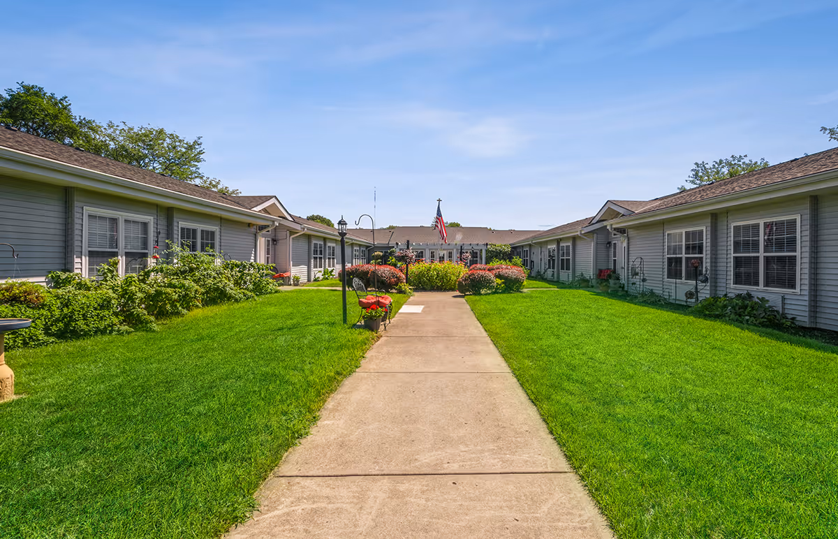 A wide concrete pathway leads through a well-maintained green lawn with bushes and flowers on either side, flanked by single-story buildings with gray siding and white-trimmed windows under a clear blue sky. An American flag is visible in the distance at the center of the buildings.