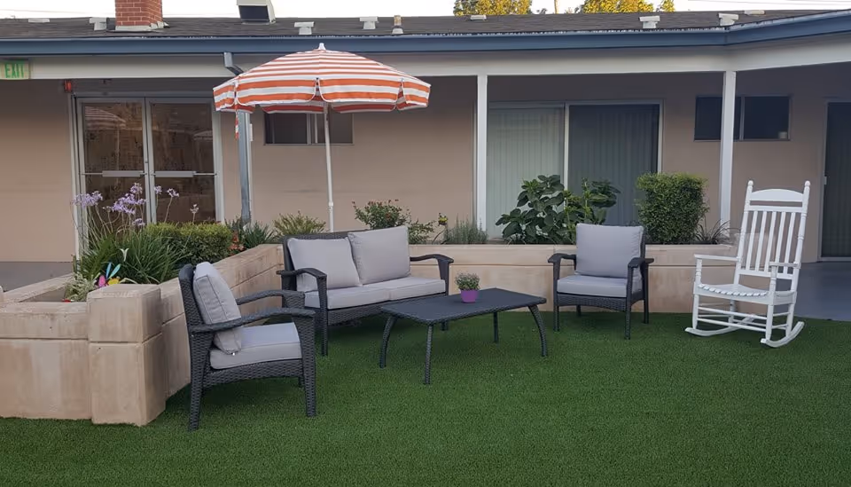 Courtyard seating area with wicker sofa and chairs, a coffee table, striped umbrella, planter boxes and a white rocking chair in front of a one-story building.