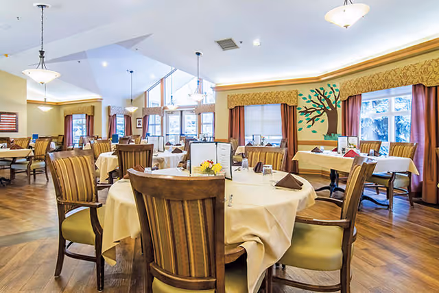 A spacious dining room with round tables covered in white tablecloths, each set with folded napkins and menus. Wooden chairs with striped cushions surround the tables. The room features large windows with curtains, a decorative tree mural on one wall, and hanging light fixtures from a high ceiling.