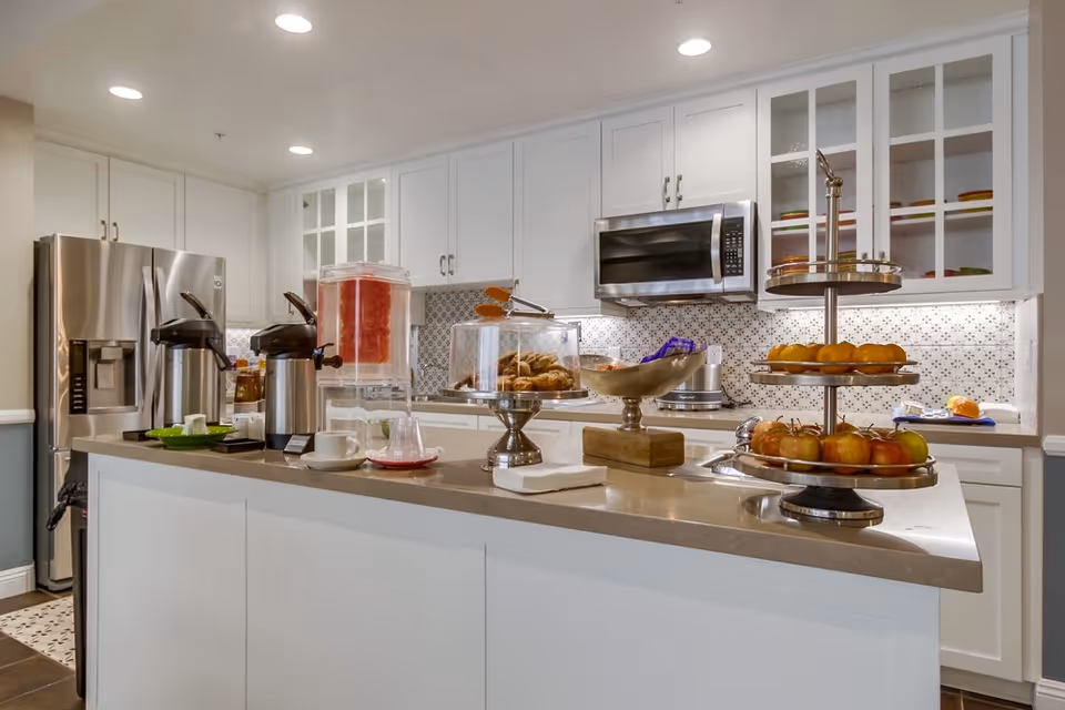 Modern kitchen area with white cabinets, stainless steel refrigerator and microwave, a countertop island with a water dispenser, coffee urns, pastries under a glass cover, a bowl of snacks, and a tiered tray holding apples and oranges.