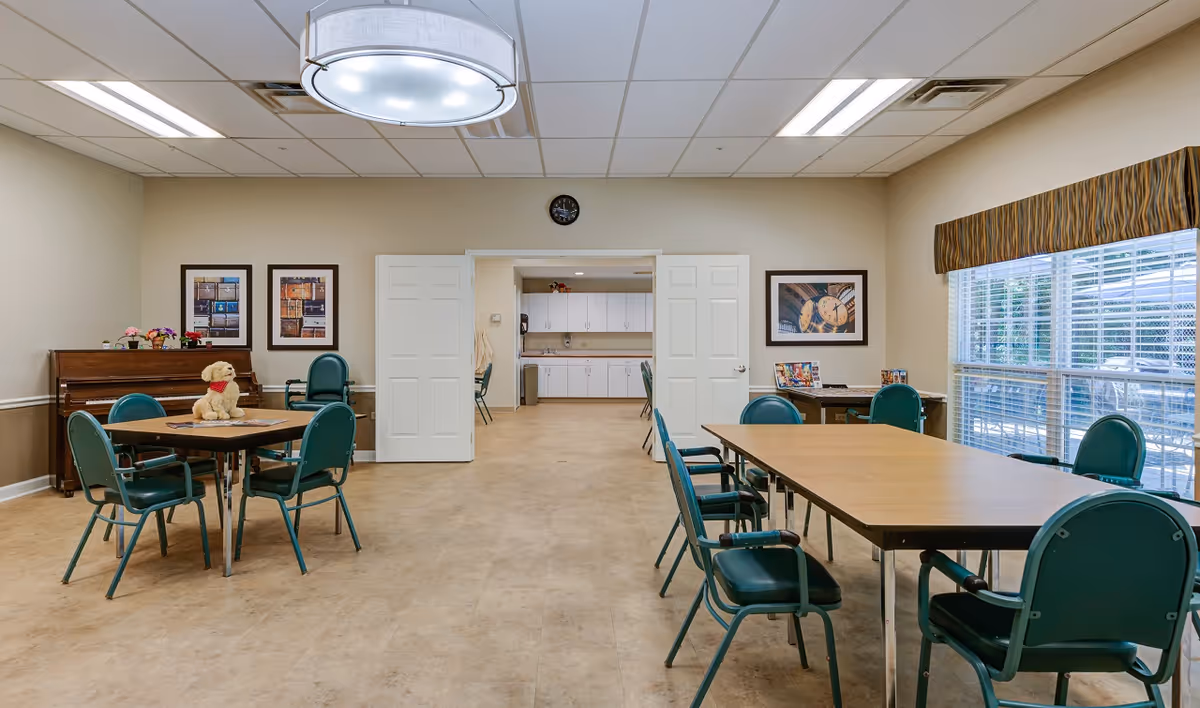 Well-lit communal dining/activity room with tables and green chairs, a piano on the left, and a kitchenette visible through double doors.