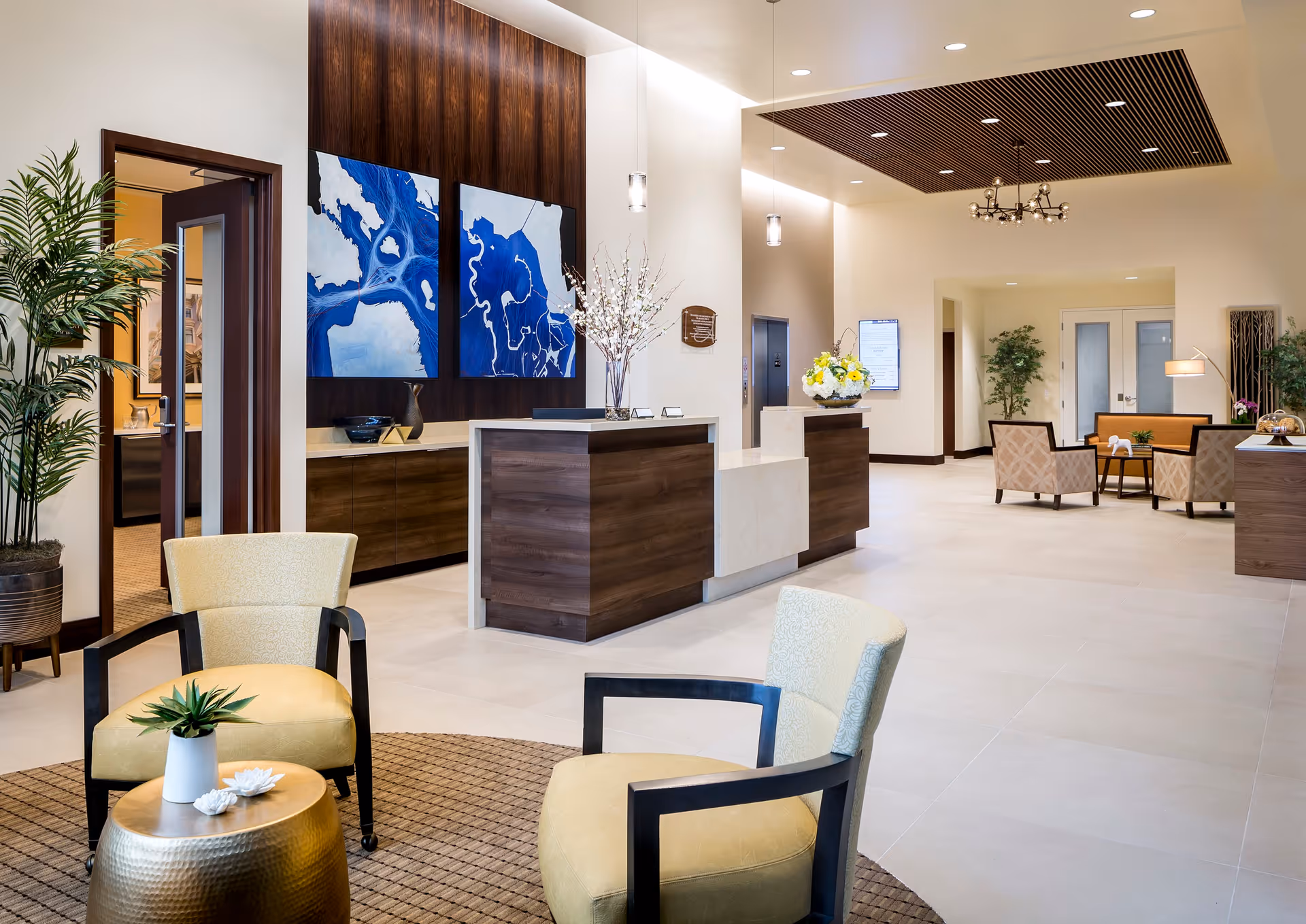 A spacious and modern senior living facility lobby with a reception desk made of wood and stone. The area features comfortable seating with yellow cushioned chairs around a small round table with a plant. There are decorative blue and white abstract paintings on the wall behind the reception desk, pendant lights hanging from the ceiling, and additional seating with armchairs and a sofa in the background. The space is well-lit with recessed lighting and has a clean, welcoming atmosphere.