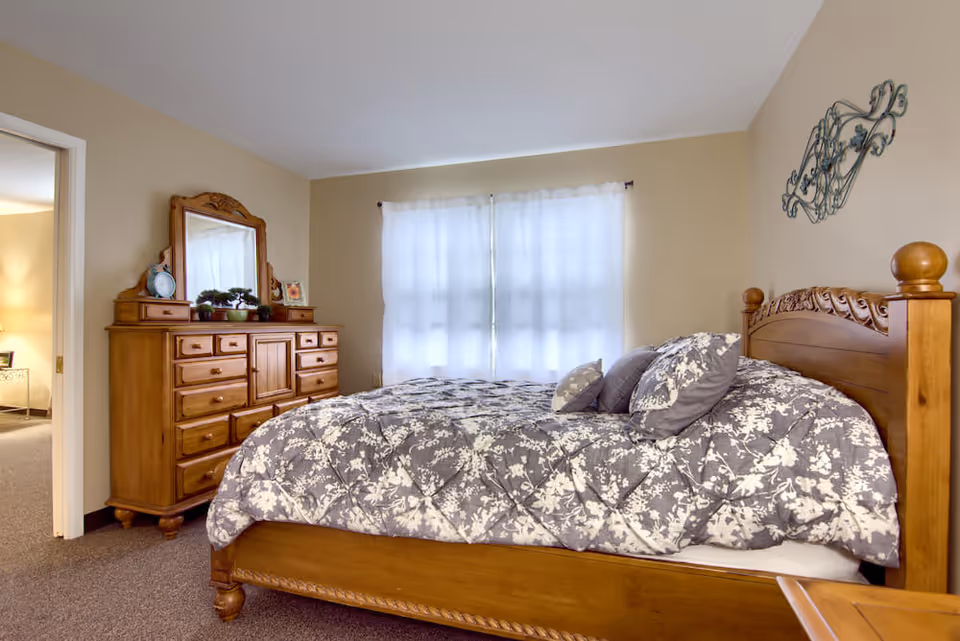 Sunlit bedroom with a wooden bed and floral bedding, matching dresser with mirror, and a curtained window.