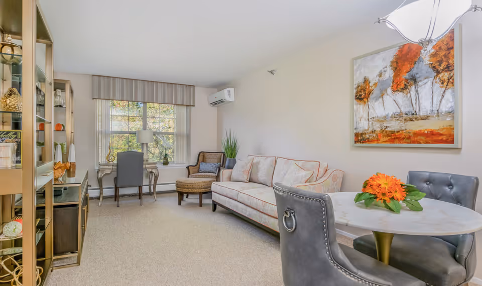Bright furnished living room with a sofa, armchairs, small round dining table, desk by a window, shelving, and wall art.