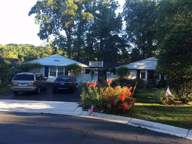 Front exterior view of a single-story residential-style building with the address number 13201 displayed. Two vehicles are parked in the driveway, and there are well-maintained bushes, flowers, and trees surrounding the property. The scene is set in daylight with clear skies.