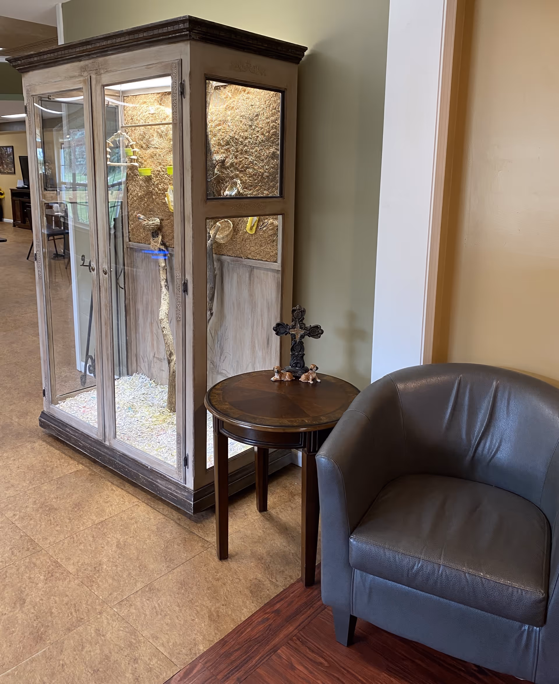 A cozy corner in an indoor space featuring a gray leather armchair next to a small round wooden table with decorative items including a cross and two small dog figurines. Behind the table is a large glass enclosure with branches and bird toys inside, likely for pet birds. The floor has a mix of wood and tile patterns, and the walls are painted in neutral tones.