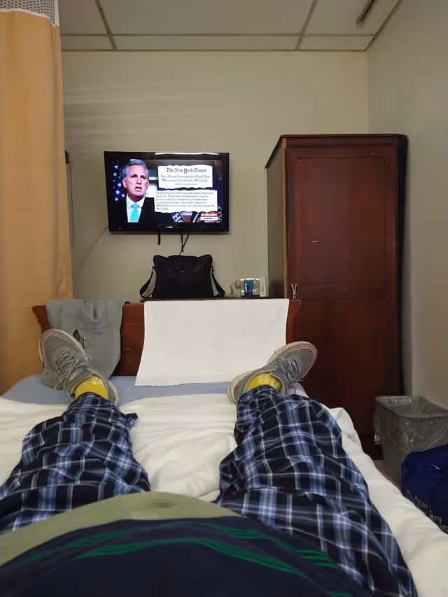View from a person lying on a bed in a room, showing their legs in plaid pajama pants and gray sneakers. A television mounted on the wall displays a news program. There is a wooden cabinet to the right, a black bag on a piece of furniture beneath the TV, and a beige curtain on the left side of the image.